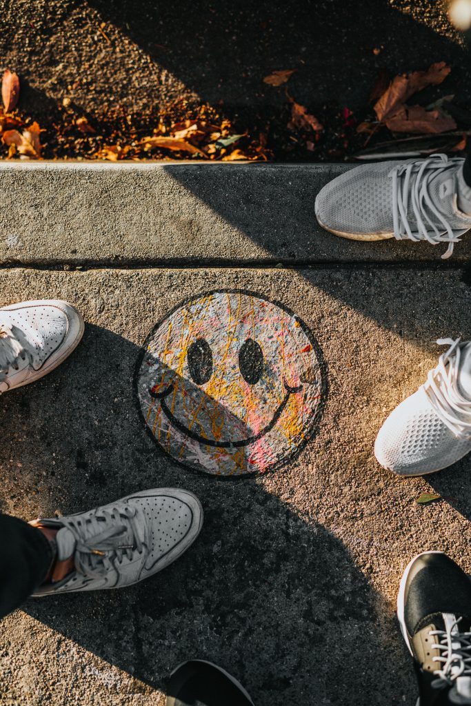 A happy smiley drawn with chalk on the street surrounded by people, only pairs of shoes visible